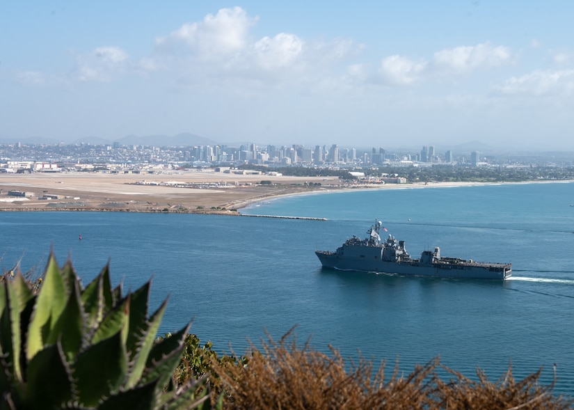 A military ship cruises at sea near a beach and a city in the distance during the daytime.