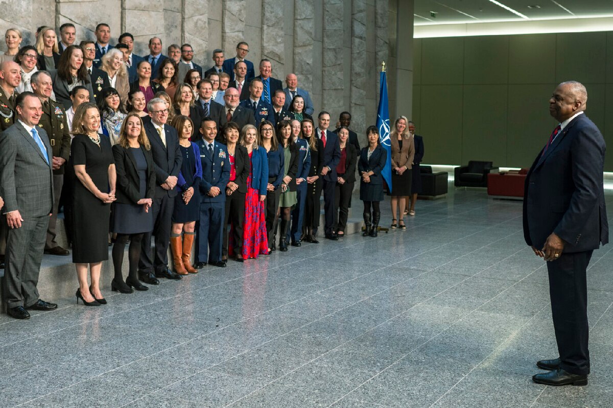 A person in business attire addresses a large group of people standing in rows.