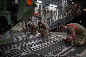 Three Airmen use chains to secure a large military vehicle inside a C-17 Globemaster III aircraft.
