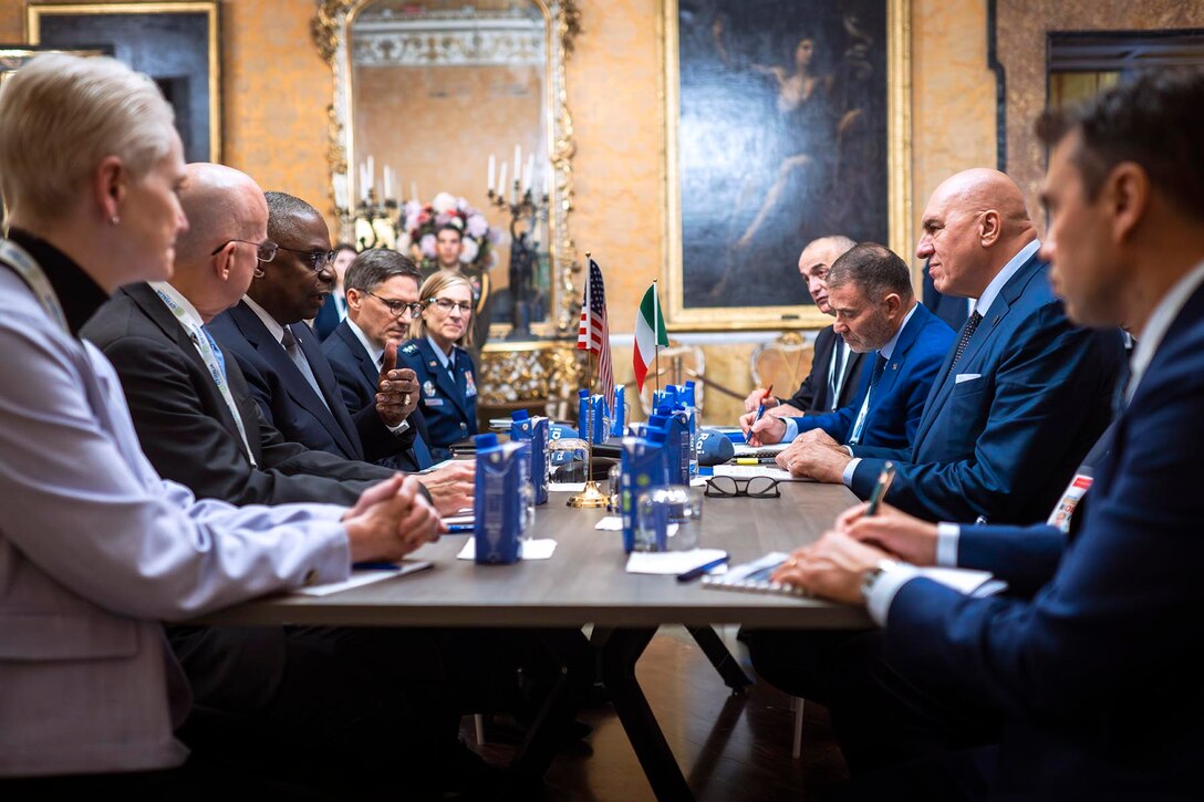 A group of government officials sit across from one another and chat. Water bottles and the American and Italy flags rest on the table.