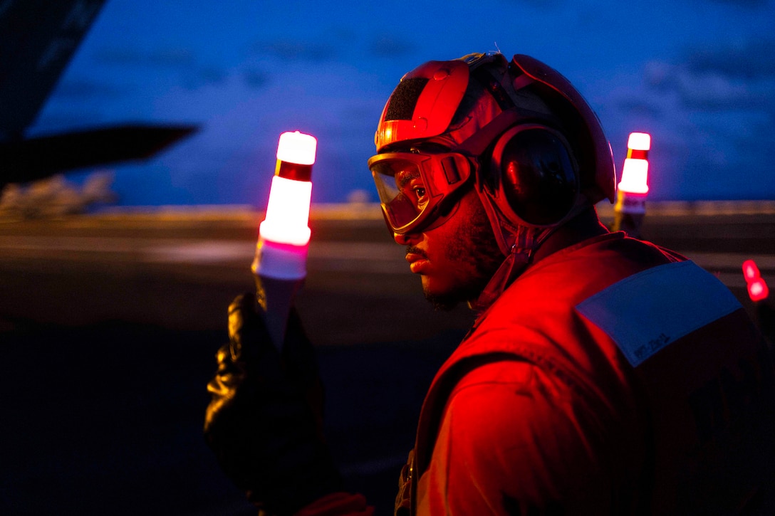 A sailor wearing safety goggles and ear protection holds two white marshalling wands while standing on an aircraft carrier at night under red light.