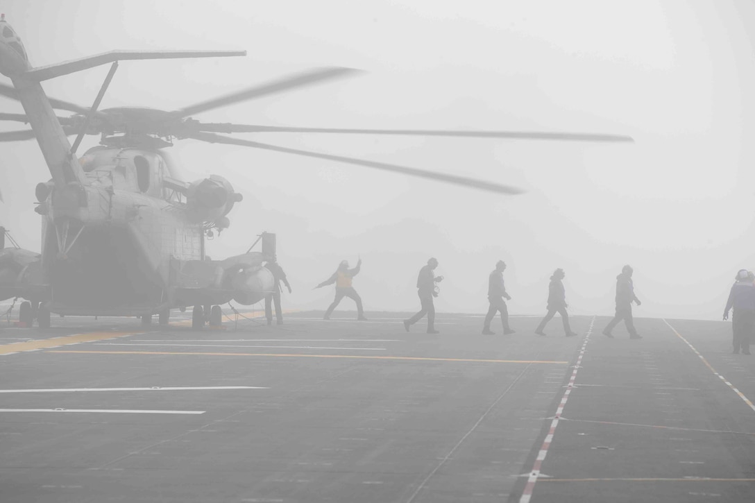 A large helicopter sits on the deck of a military ship in the fog as service members walk away from it.