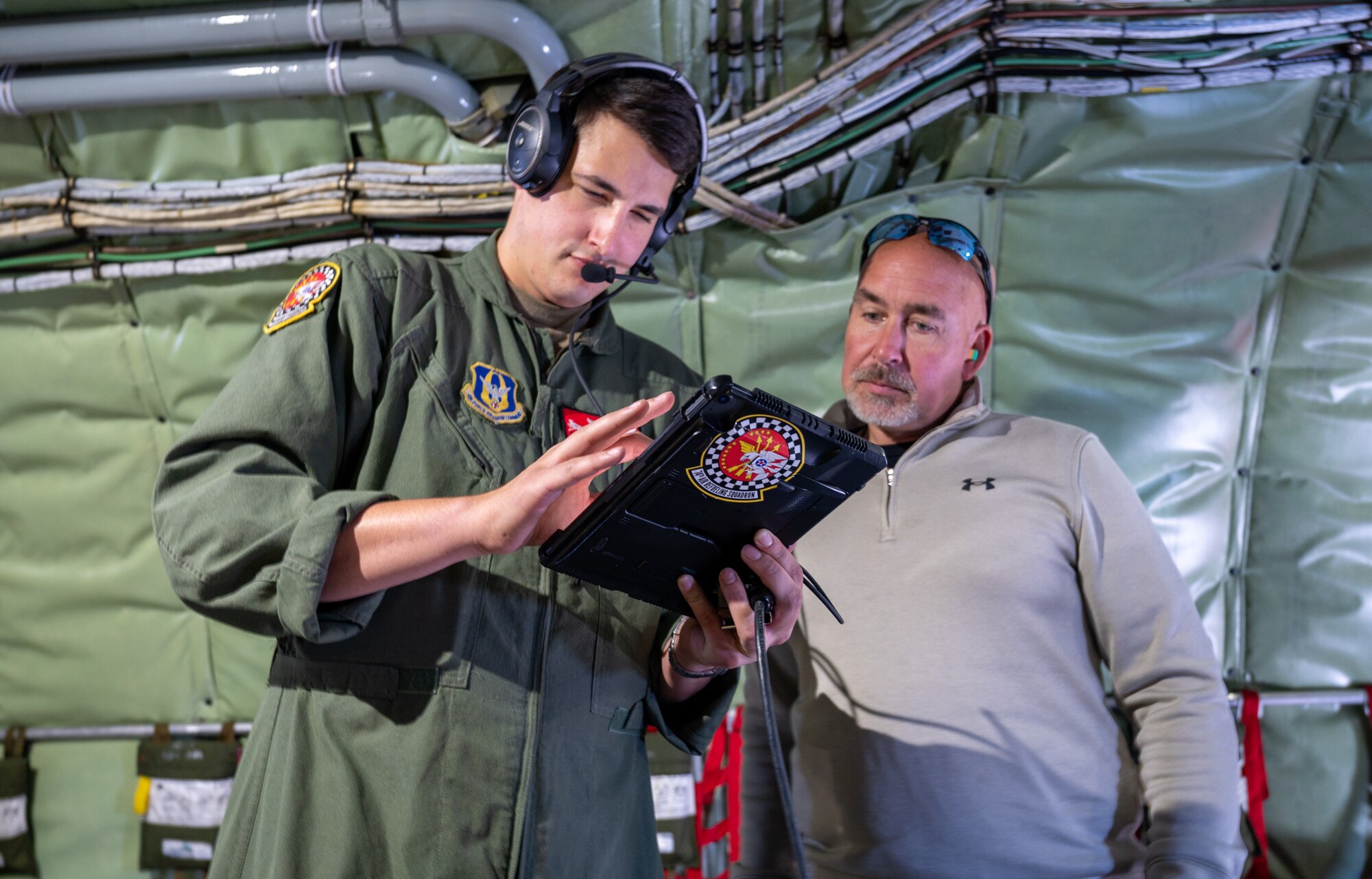 A man wearing a flight suit gestures to his tablet screen while talking to a civilian while aboard a military aircraft. Both men are standing.
