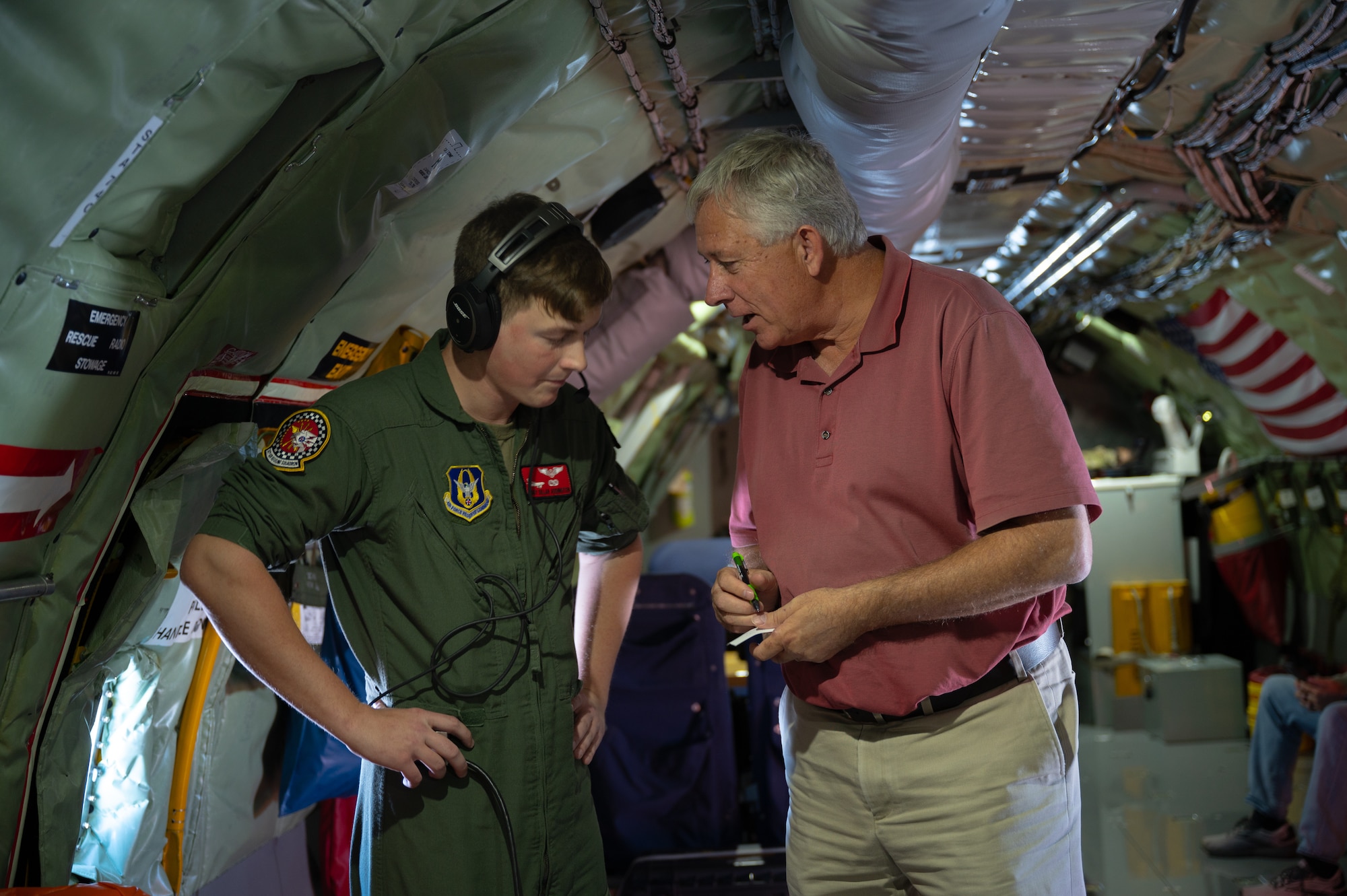 Two men standing in a military aircraft have a conversation. The man at left is in a flight suit while the other is in civilian clothing.