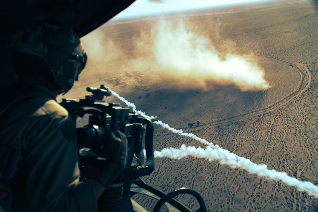 A Marine fires a weapon from an airborne aircraft leaving a streak a smoke in the sky as smoke blows from a desert-like area below.