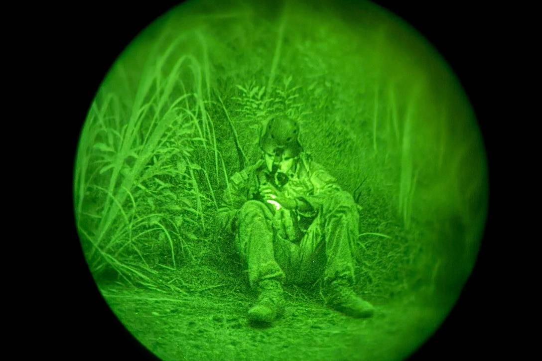 A soldier in combat gear sits in tall grass as seen through night vision lens.