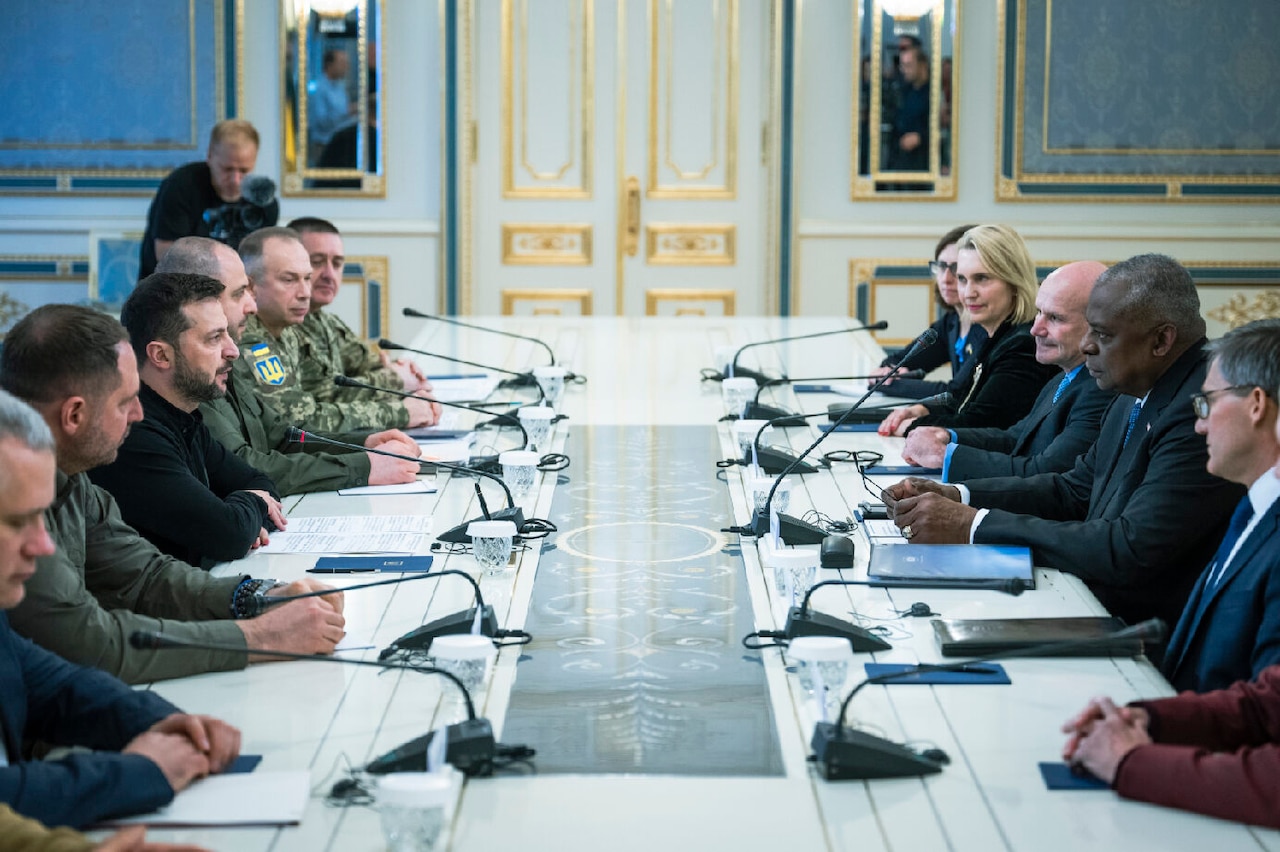 A group of people wearing business attire and foreign military uniforms sit around a table with microphones.