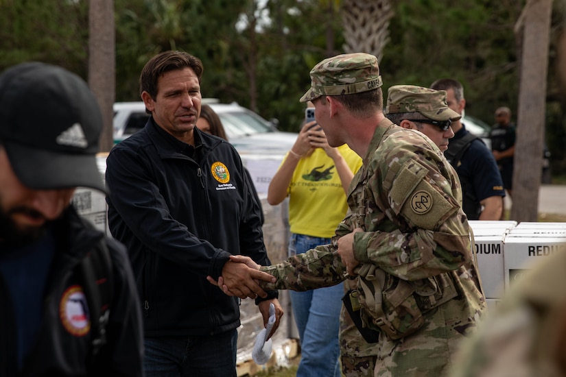 A government official in a sweater shakes hands with a guardsmen outside during the day.