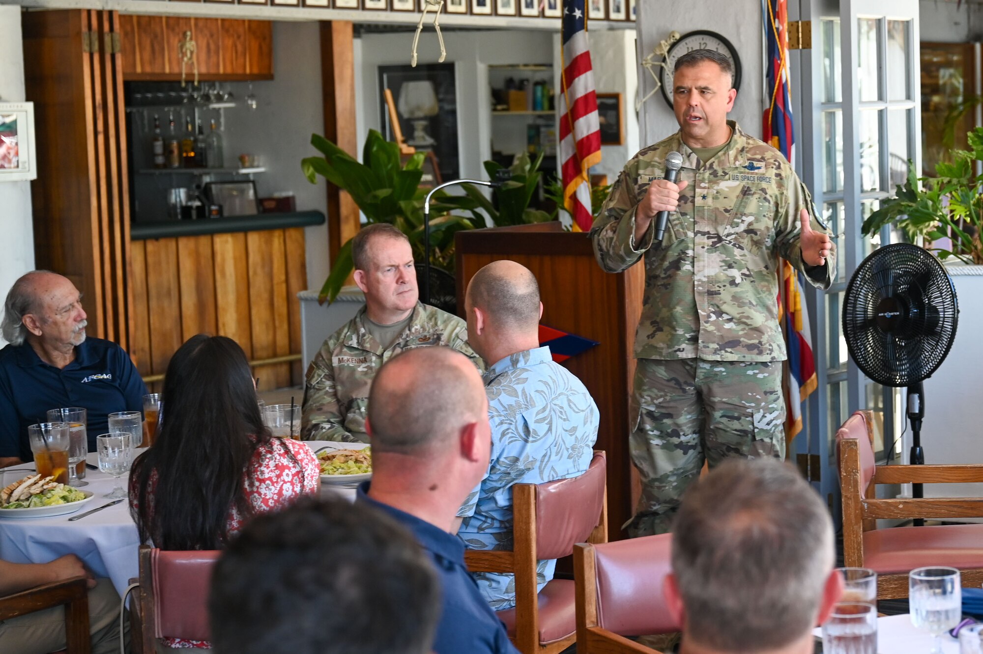 A man in military uniform stands in front a dinning area and speaks into a microphone