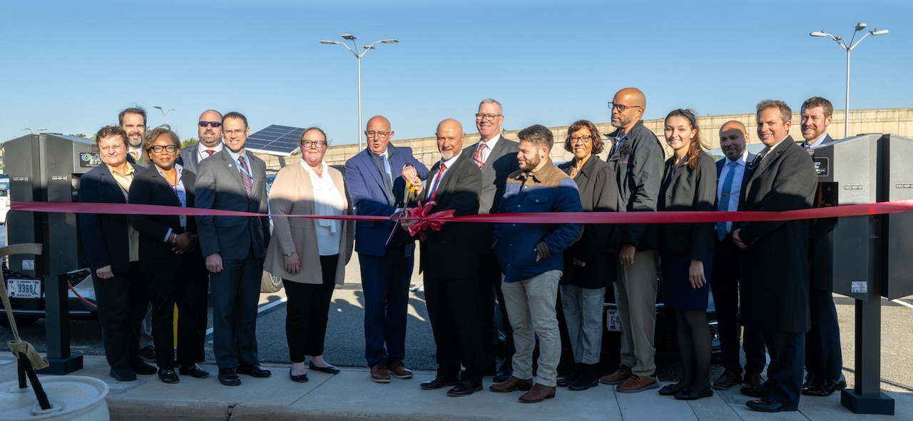 Men and women stand behind a ribbon. One man holds a large pair of scissors and prepares to cut the ribbon.