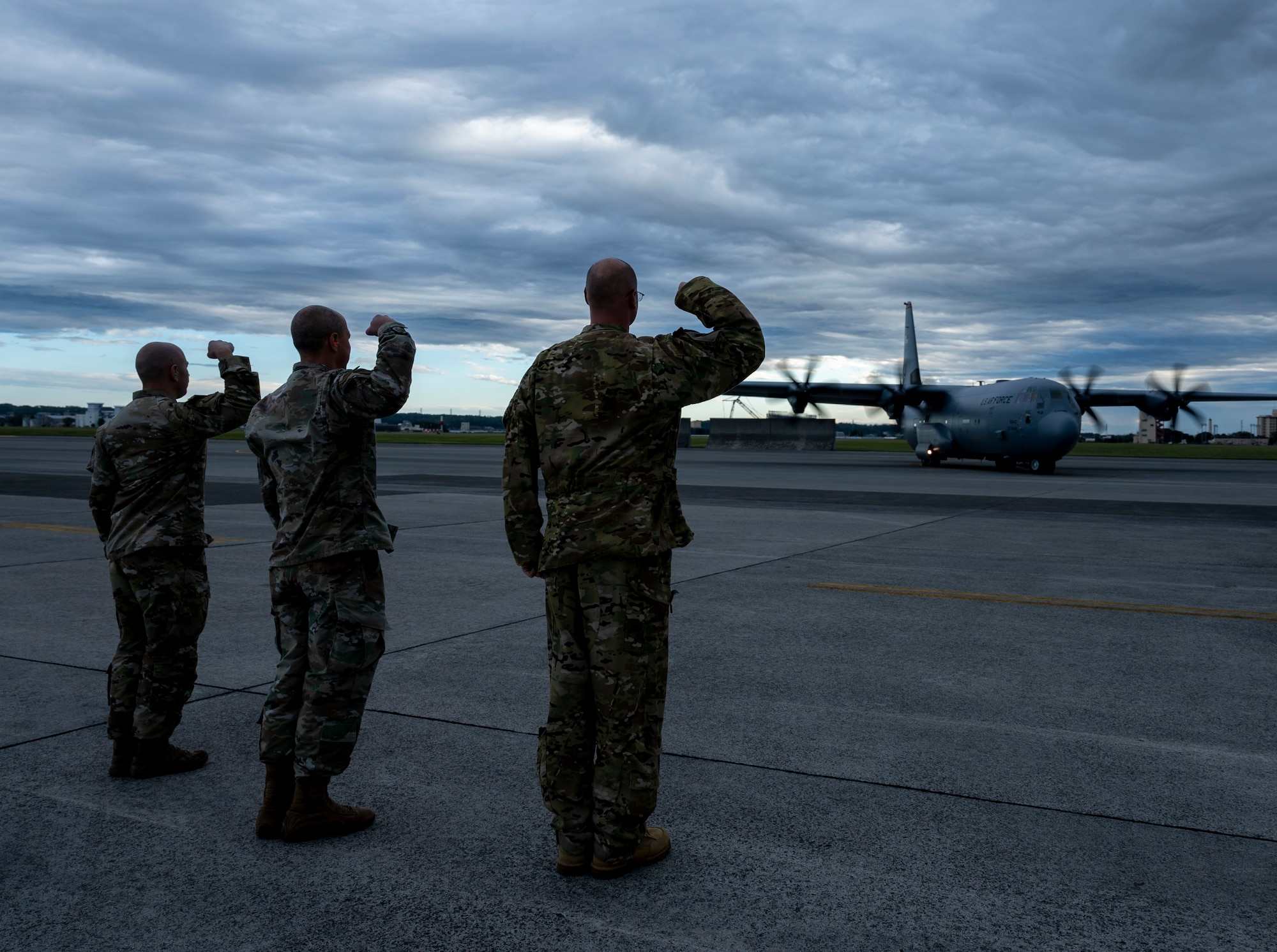 Three military members hold up their arms at an angle has a C-130J aircraft taxis the flightline.