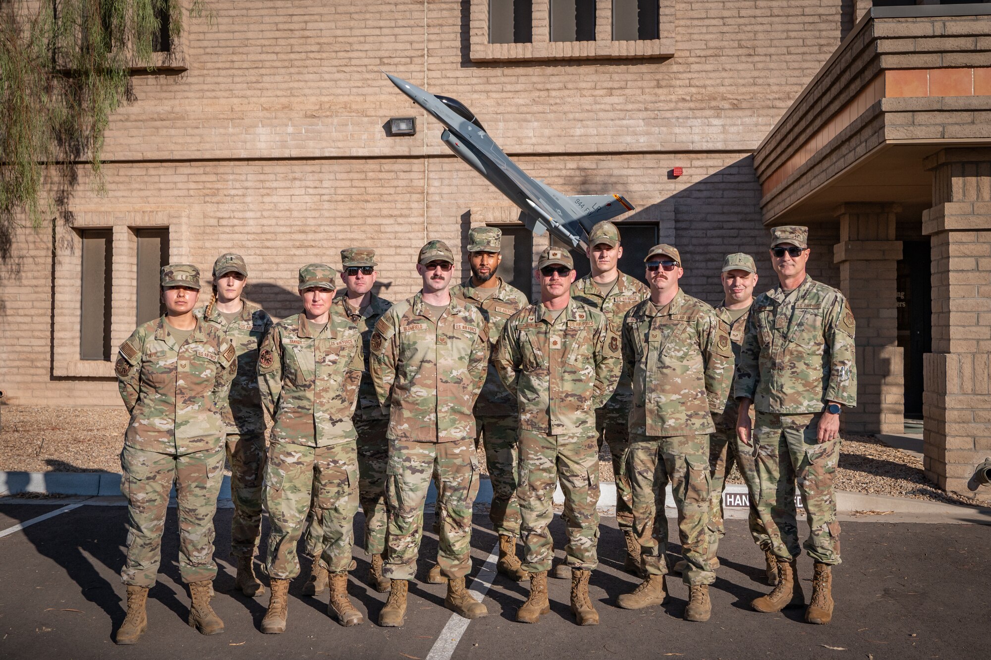 944th Fighter Wing maintainers stand for a group photo with the newly installed F-16 static display outside wing headquarters at Luke Air Force Base, Ariz., Oct. 17, 2024. The nearly quarter-scale F-16 now stands as a symbol of the wing's legacy and a tribute to those who have served, as outlined in the story "Hellion Reborn: Restored F-16 Display Honors 944th FW Legacy Future." (U.S. Air Force photo by Tech. Sgt. Tyler J. Bolken)