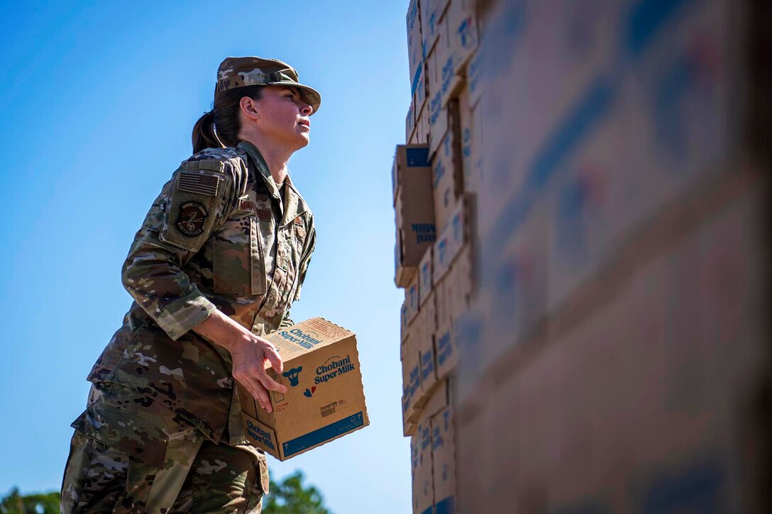 An airman carries a box during a clear blue sky next to a pile of boxes.