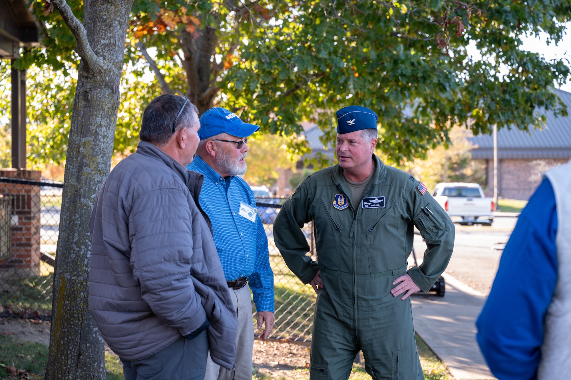 Three men stand outside in a semi-circle, having a conversation. The man at right is wearing a U.S. Air Force flight uniform while the other two men are dressed in civilian attire. The arm of another civilian is barely visible at the far right edge of the frame.