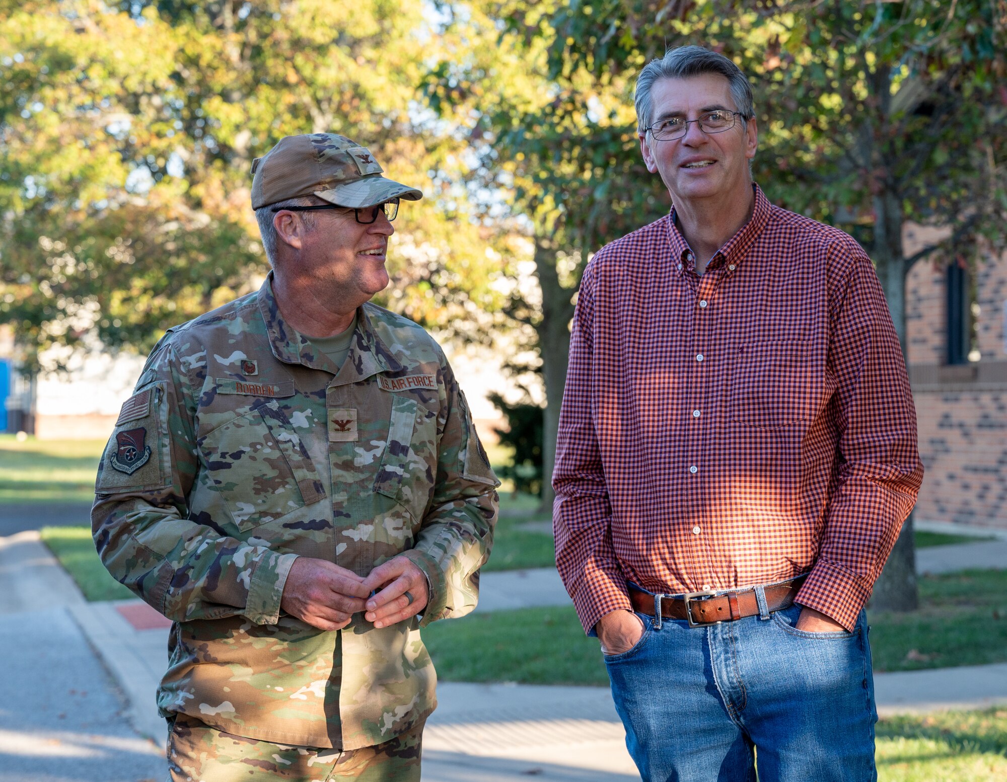 Two men stand alongside each other outside on a fall day. The man on the left is dressed in an Air Force camouflage uniform, the other dressed in jeans and a dress shirt.
