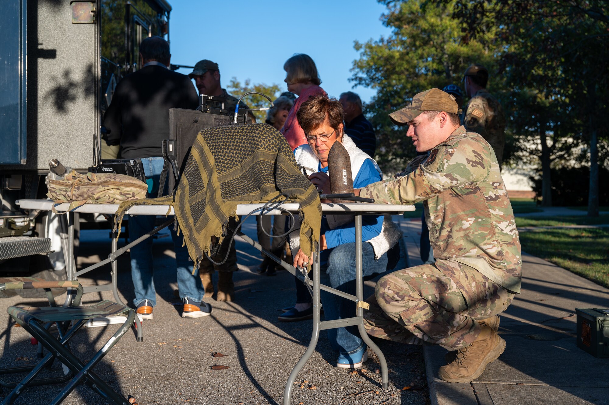 A uniformed military members and civilian woman are crouched down, looking at a computer on a table outside on a fall day. In the background, silhouettes of civilians are visible.