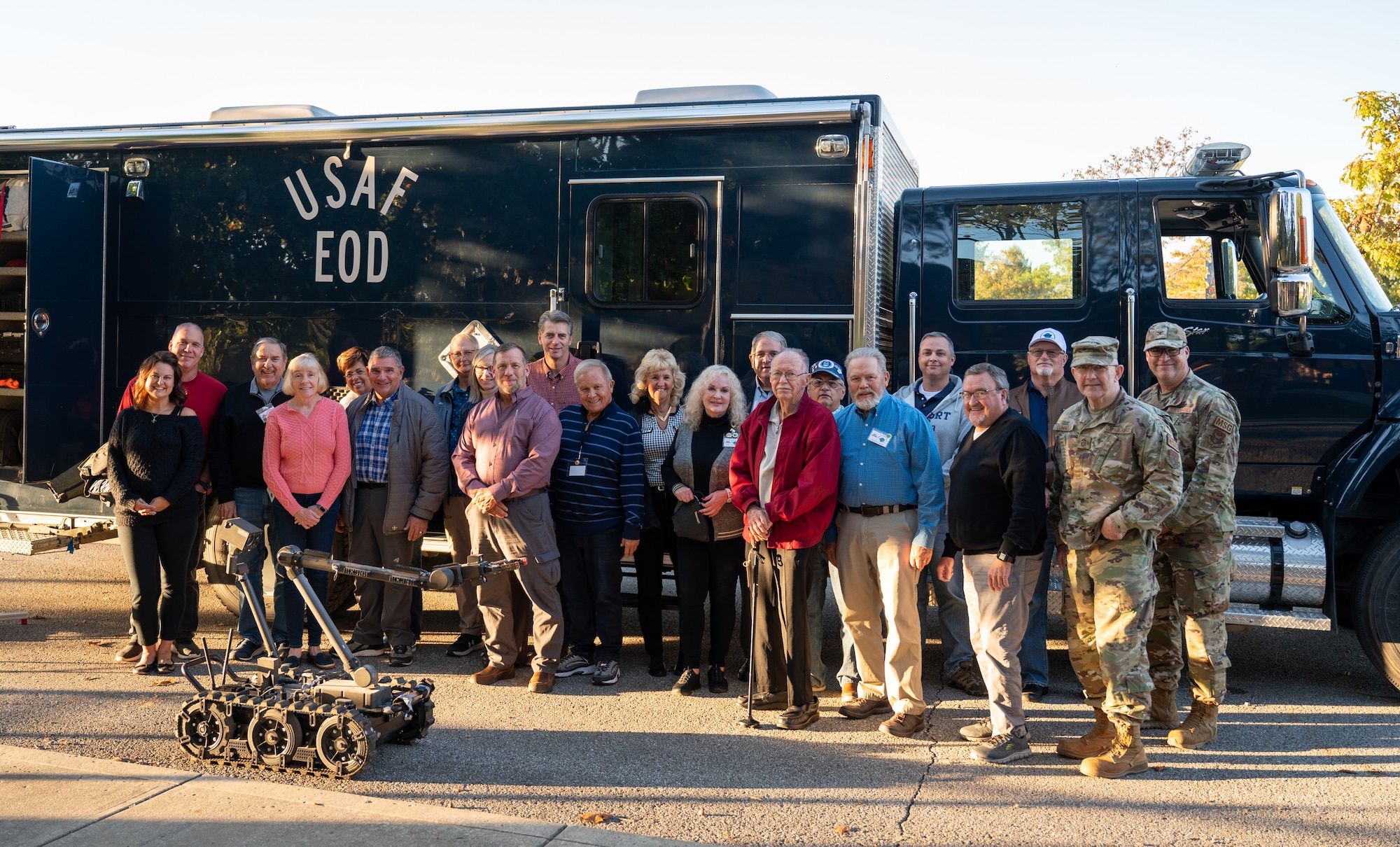 A group of people stand in front of a large blue truck that reads "USAF EOD", posing for a group photo outside on a fall day.