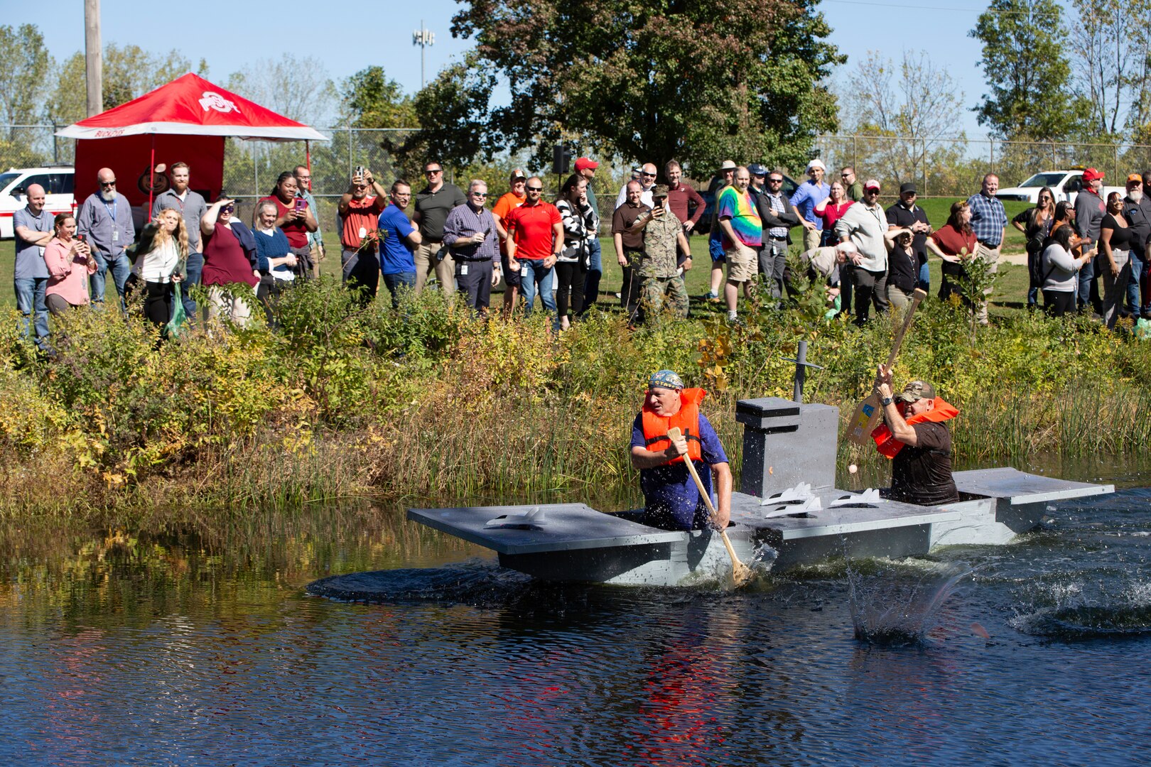 A cardboard boat shaped like an aircraft carrier makes its way down the length of a pond. Two men paddle it.