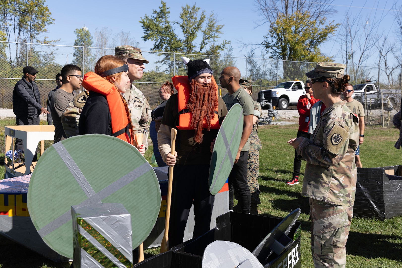 A group of people chat near a cardboard boat on land. Two are dressed like Vikings (woman and man) and two are in military uniforms, one is a woman and the other a man.