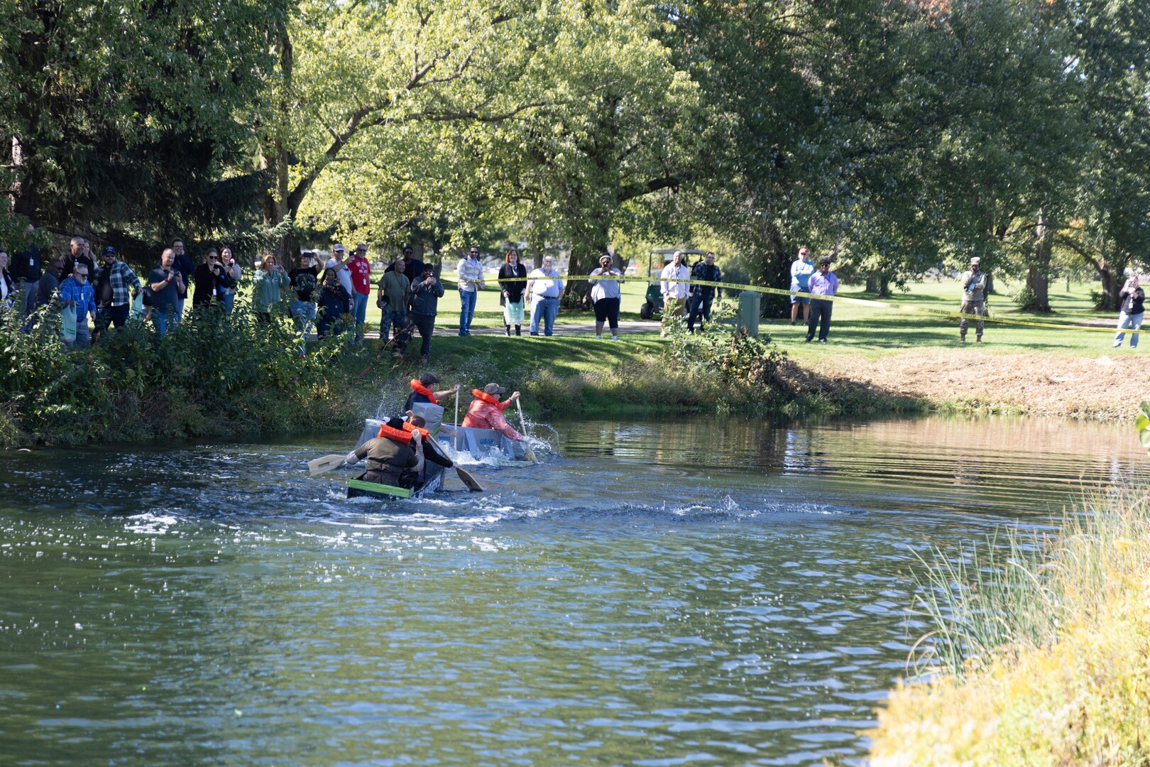 Two cardboard boats splash around in a pond outside with trees and people watching in the background.