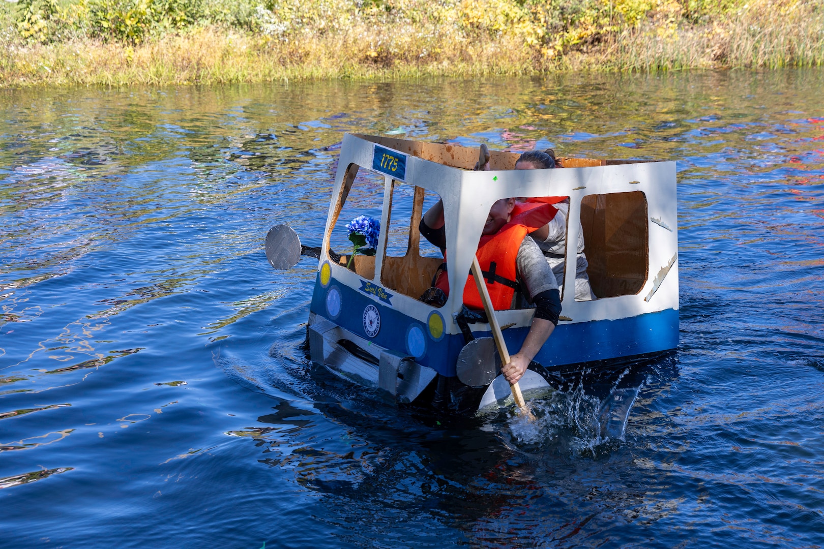 A small bus shaped boat makes its way down a pond. It is paddled by two women.