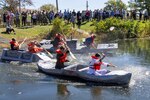 Cardboard boats start a race down the length of a pond. Several people are paddling each boat.
