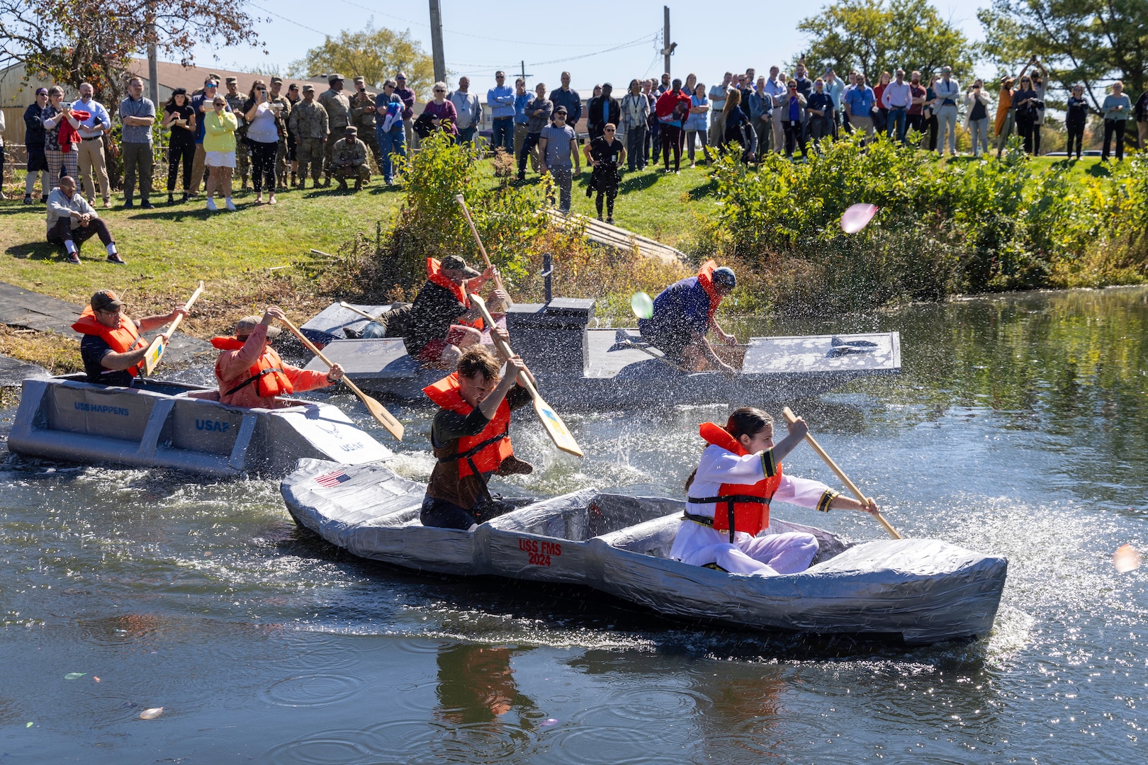 Cardboard boats start a race down the length of a pond. Several people are paddling each boat.
