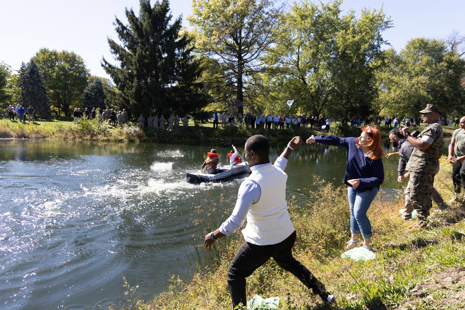 A cardboard boat shaped makes its way down the length of a pond. Two people paddle it. People are throwing water balloons at it as well.