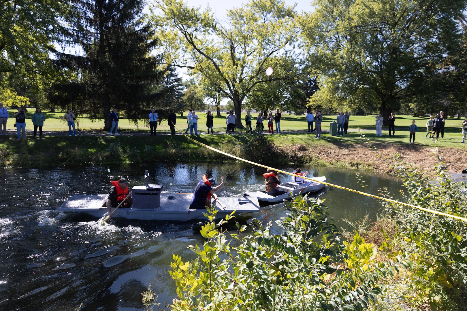 Two cardboard boats fight at the finish in a pond. Four people are paddling.