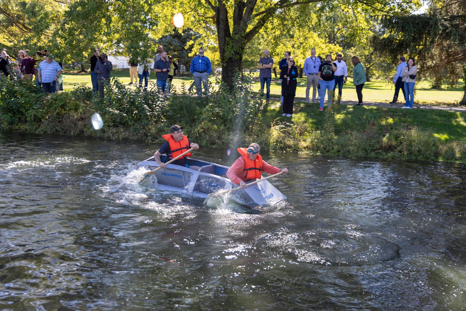 A cardboard boat shaped like bullet makes its way down the length of a pond. Two men paddle it.