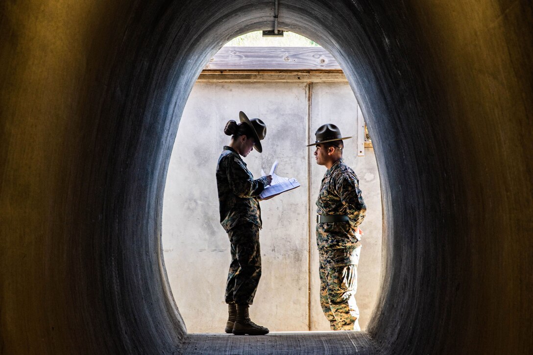 Two Marines stand facing each other, one looking at papers, framed by a tunnel-like opening in an outdoor structure.