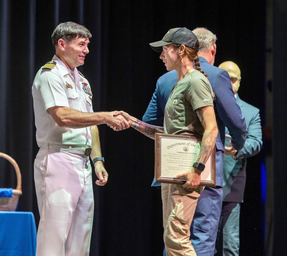 Capt. JD Crinklaw, commander, Puget Sound Naval Shipyard & Intermediate Maintenance Facility, congratulates Maria Gibson, marine pipefitter, Shop 56, Pipefitters, as she receives her apprentice graduation certificate during the 2024 PSNS & IMF Apprentice Program graduation ceremony, Sept. 20, 2024, at Bremerton High School, in Bremerton, Washington. (U.S. Navy photo by Wendy Hallmark)