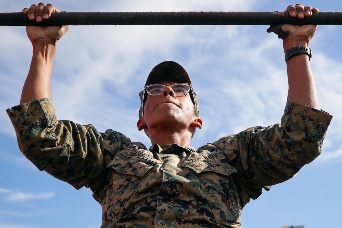 A close-up of a sailor doing a pullup under a cloudy, blue sky.