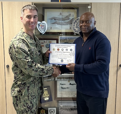 Navy Cmdr. John O’Donnell shakes hands while holding a certificate with Gerol Meadows in front of a display case