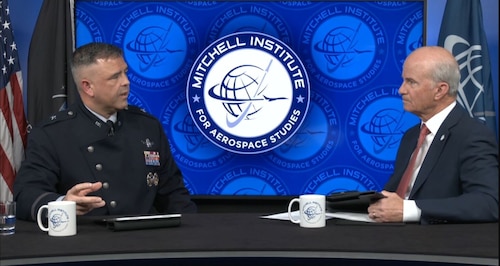 A man in a dress military uniform and a man in civilian business attire are seated across from each other at a desk, having a conversation. Behind them on the wall is a logo that reads “Mitchell Institute for Aerospace Studies.”