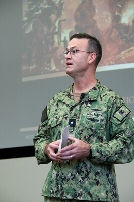 A Navy Officer in a camouflage uniform stands on an indoor stage speaking to a crowd.