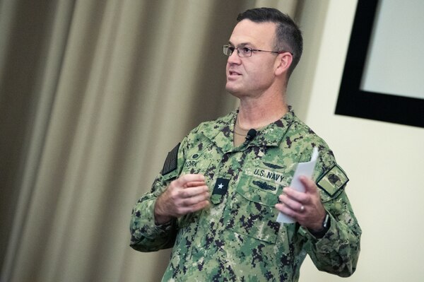 A Navy Officer in a camouflage uniform stands on an indoor stage speaking to a crowd.