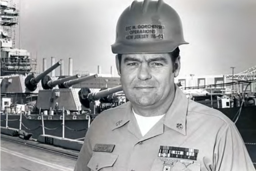 A sailor stands in front of a docked battleship.