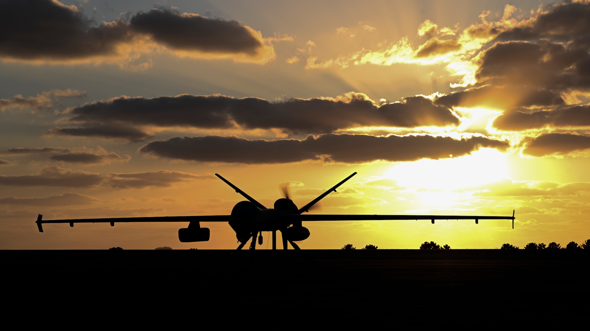 An MQ-9 unmanned aerial vehicle sits on a runway, silhouetted at dusk with a bring setting sun in the background.