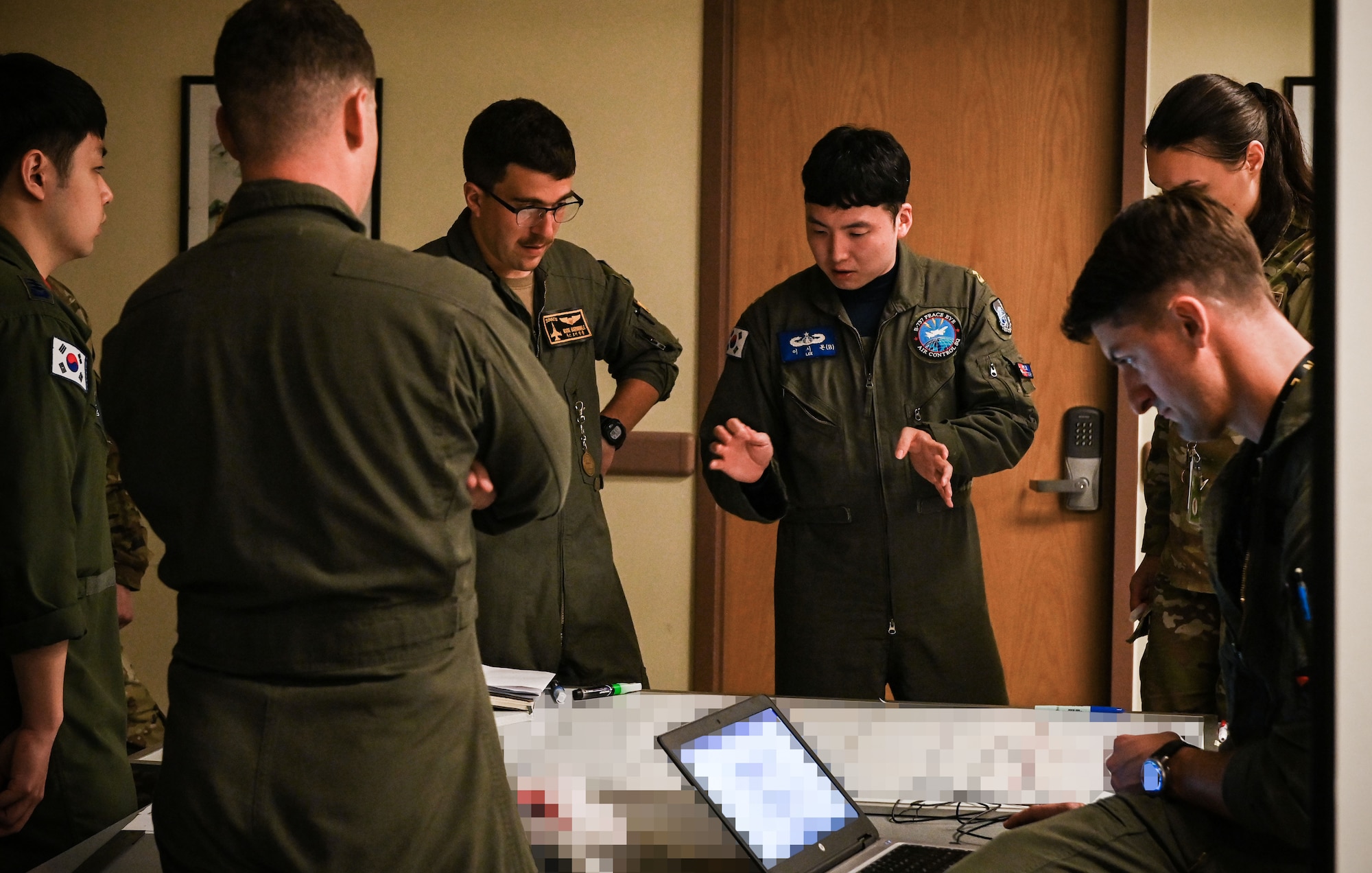 A group of 5 men in flight suits and one woman in military camouflage stand around a table with papers on it, some with their backs to the camera.