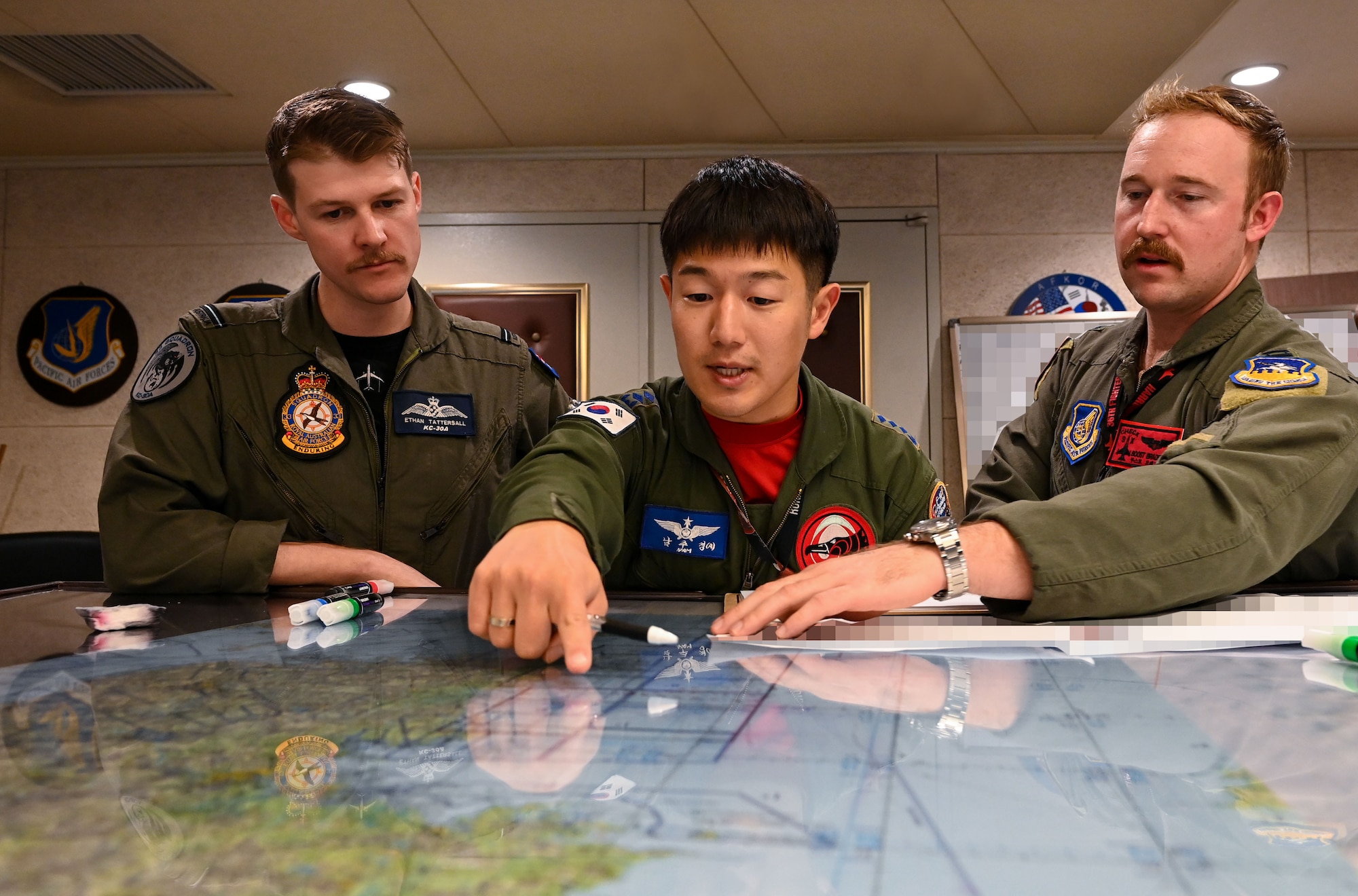 Three men in flight suits stand behind a chest-high table with maps on it, as the man in the middle points at a map on the table.