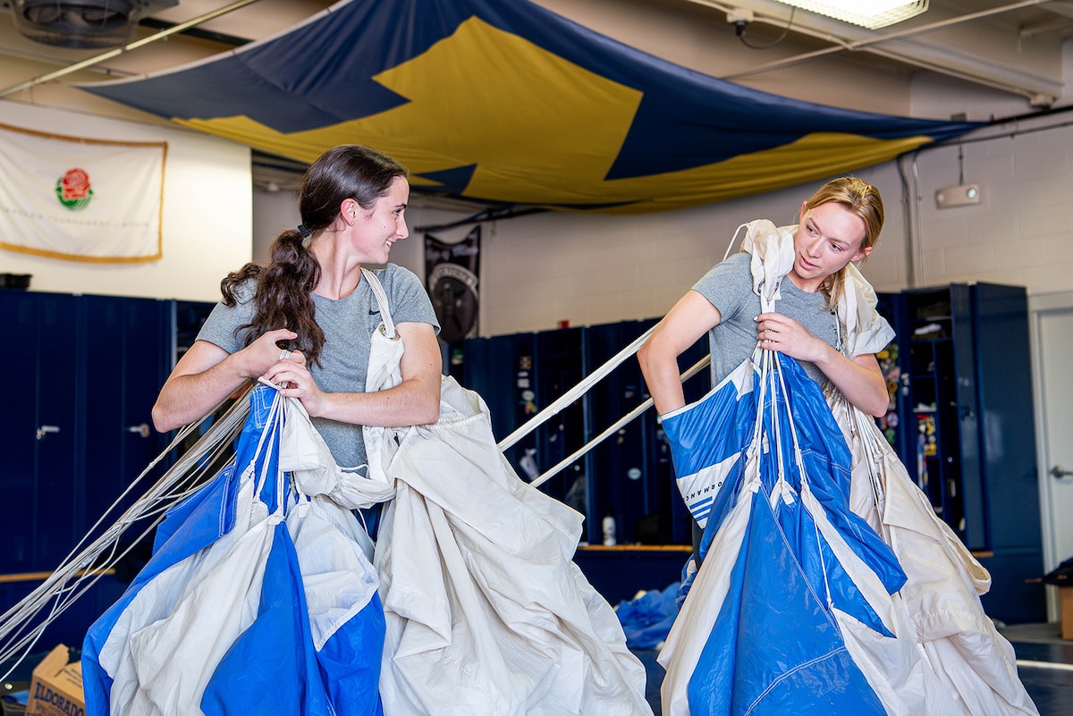 Two U.S. Air Force Academy cadets look at each other while carrying large parachutes over their shoulders inside a locker room.