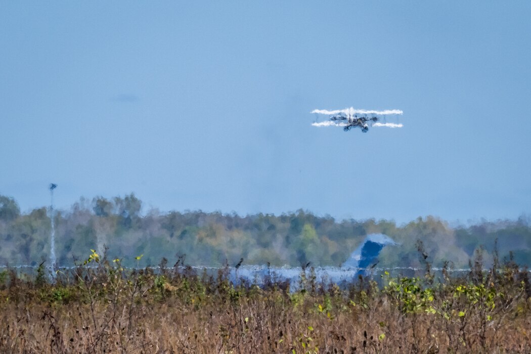 A replica of the Wright Flyer soars in the distance, distorted by heat waves, with a C-17 parked below