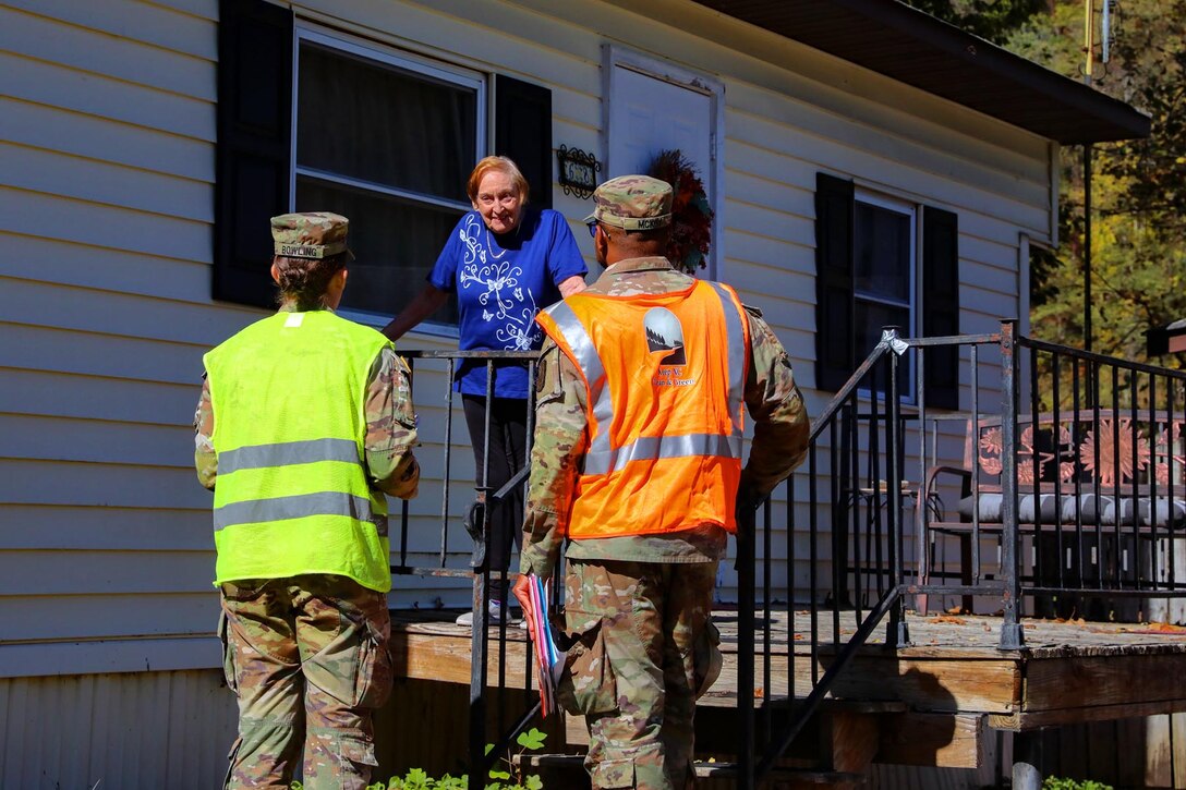 A couple of guardsmen wearing neon vests chat with a resident on her porch outside during the day.