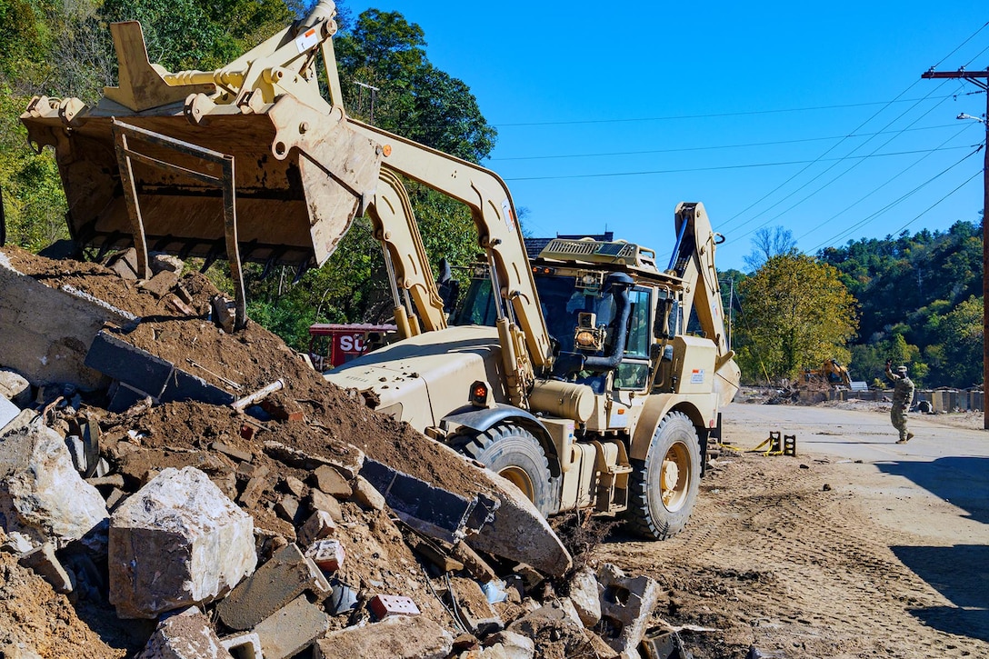 An excavator moves dirt and rock from a road to a pile of dirt and rock, while a soldier looks on from the road during the day.