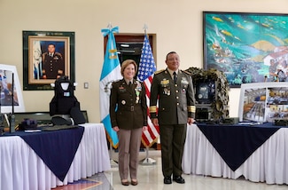 Two military personnel stand and pose for a photo in front of flags.