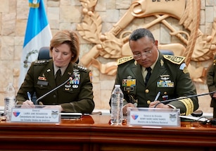 Uniformed military personnel sign an agreement at a table.