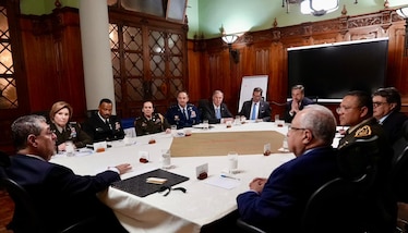 Uniformed military personnel and a man in a suit talk at a conference table.