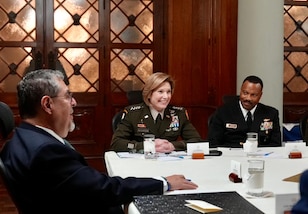 Uniformed military personnel and a man in a suit talk at a conference table.