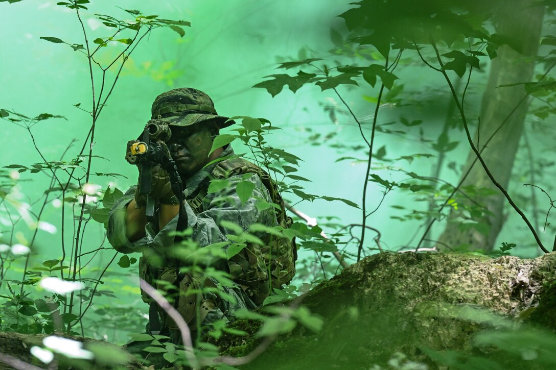 An airman points a weapon while kneeling amid foliage and trees with green smoke permeating the air.